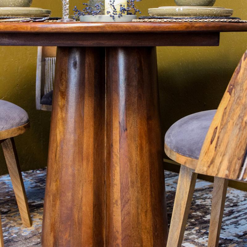 A close-up of a rustic wooden dining table with a thick, rounded pedestal base, surrounded by matching wooden chairs with gray upholstered seats.