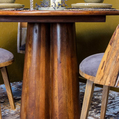 A close-up of a rustic wooden dining table with a thick, rounded pedestal base, surrounded by matching wooden chairs with gray upholstered seats.