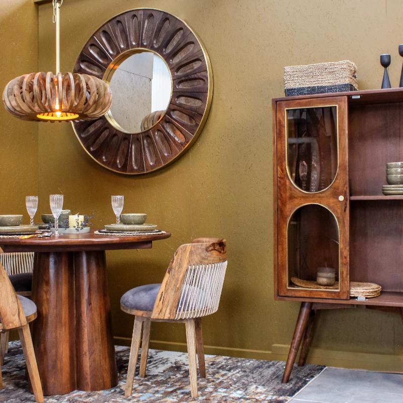 A mid-century modern dining room scene featuring a round wooden table, woven-back chairs, a dark wood cabinet with glass doors, a large sunburst wall mirror, and a textured olive green wall.