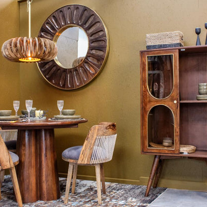 A mid-century modern dining room scene featuring a round wooden table, woven-back chairs, a dark wood cabinet with glass doors, a large sunburst wall mirror, and a textured olive green wall.