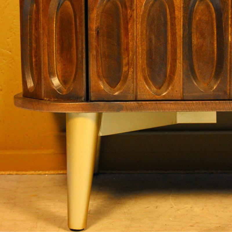 Close-up of a mid-century modern wooden cabinet with carved oval door panels and tapered cream-colored legs, shown in a warm indoor setting.