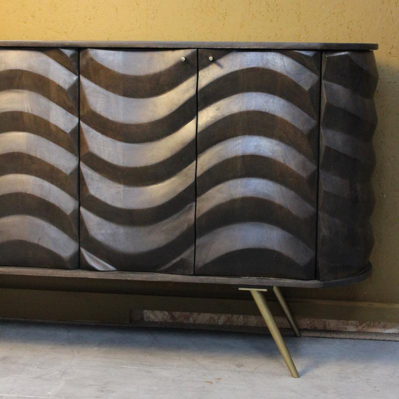 A mid-century modern sideboard cabinet with three doors featuring a wavy, textured metallic pattern in silver and dark bronze tones, supported by slim gold-toned legs against a textured yellow wall.