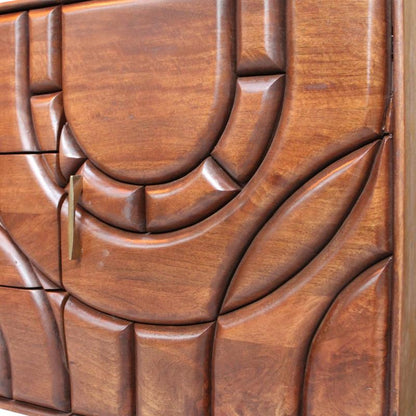 Mid-century modern wooden cabinet with sculptural curved panel detailing and a brass pull handle, photographed in an indoor setting.