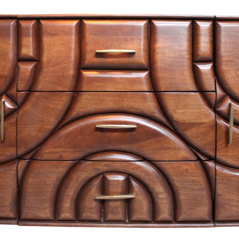 A close-up of a mid-century modern wooden sideboard or dresser with sculpted geometric drawer fronts and curved arch details, featuring brass handles against a warm beige wall.