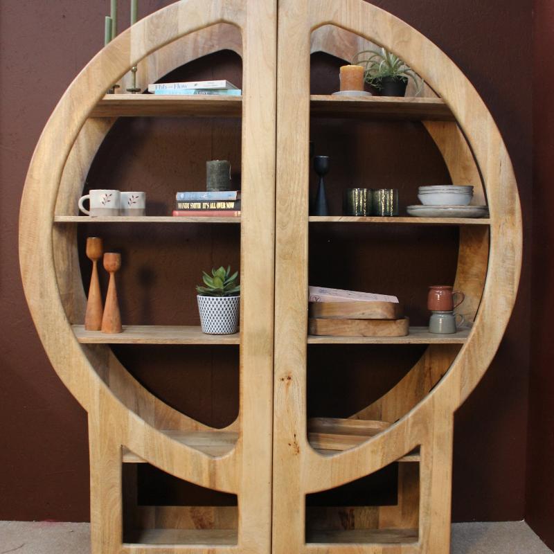 A large round wooden display cabinet with two doors and multiple open shelves, styled with books, cups, small plants, and decorative objects against a dark brown wall.