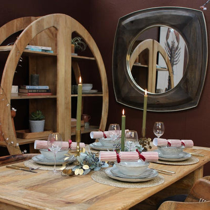 A rustic wooden dining table set for four with plates, wine glasses, striped napkins, and tall green candles, in a styled dining room with a wooden hutch, wall mirror, and woven chair.