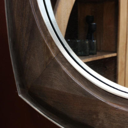 Close-up of a round wall mirror with a dark wood frame and a silver metal trim, mounted on a dark reddish-brown wall. The reflection shows wooden shelves and a candle.