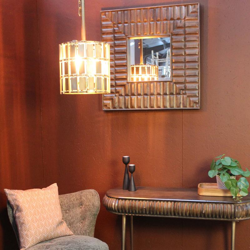 A styled interior corner with a gray upholstered accent chair, a round dark wood coffee table, a narrow console table, a patterned rug, a hanging woven pendant light, and a rectangular woven wall mirror against a warm rust-brown wall.