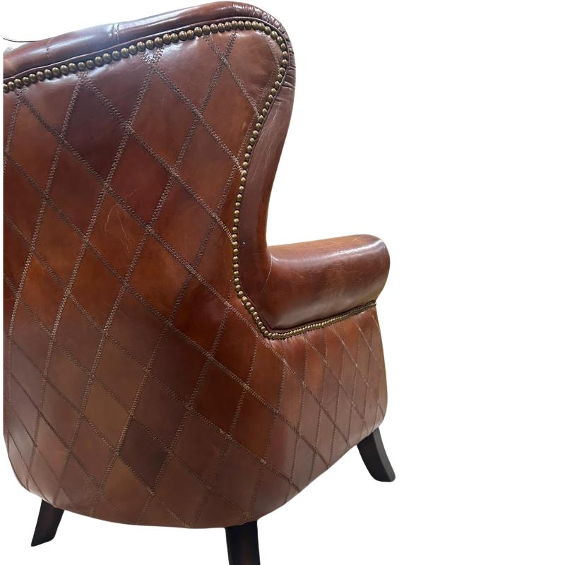 A close-up of a brown leather wingback armchair showing the quilted diamond pattern, brass nailhead trim, and dark wooden legs against a white background.