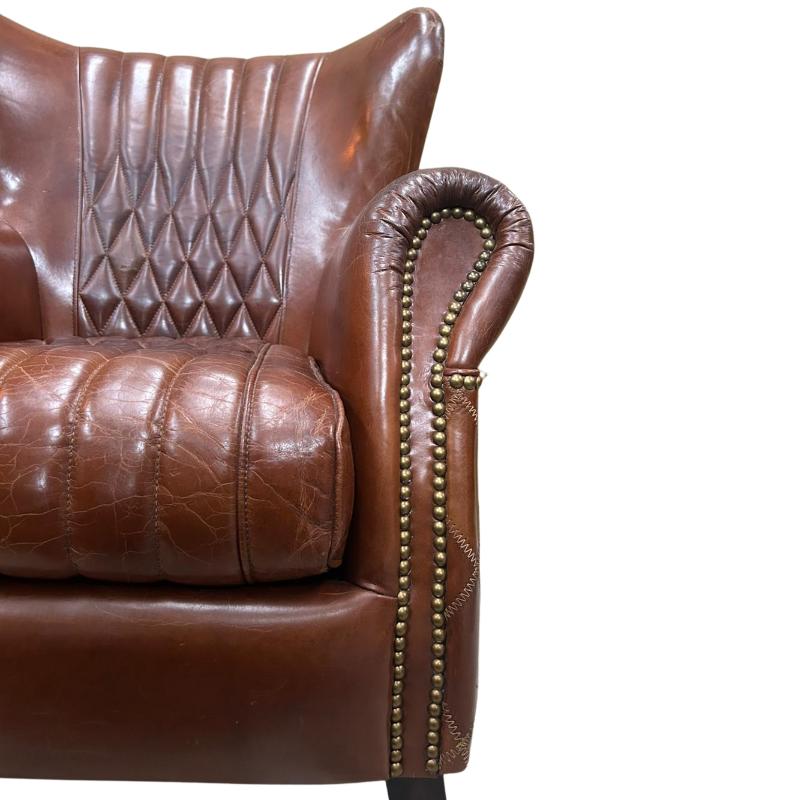 Close-up of a brown leather wingback armchair showing the quilted backrest, cushion, and rolled arm with brass nailhead trim against a white background.