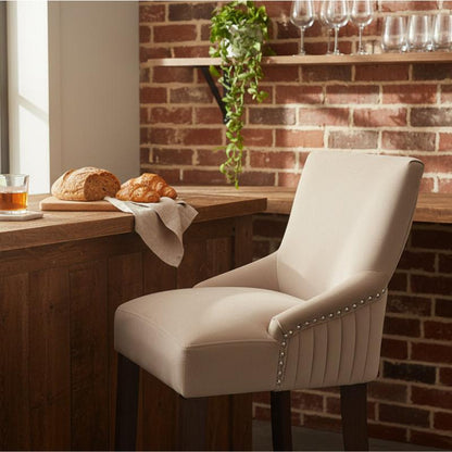 A cream upholstered barstool with dark wooden legs and silver nailhead trim, positioned in front of a rustic wooden counter in a warm, sunlit room with a brick wall background.