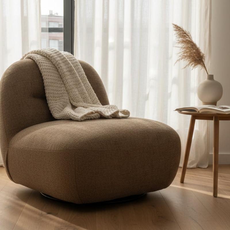 A modern low-profile lounge chair upholstered in textured taupe-brown fabric, styled with a chunky cream knit throw blanket. The chair is set in a bright, sunlit room with sheer white curtains, a light wood floor, and a small wooden side table holding a vase with dried grass and an open book.