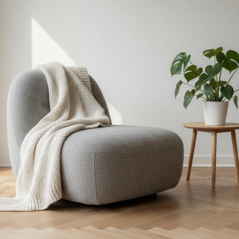A modern low-profile lounge chair upholstered in light gray fabric, styled with a cream knit throw blanket draped over the seat. The chair is set in a minimalist room with light wood flooring, a white wall, and a small wooden side table holding a potted green plant.