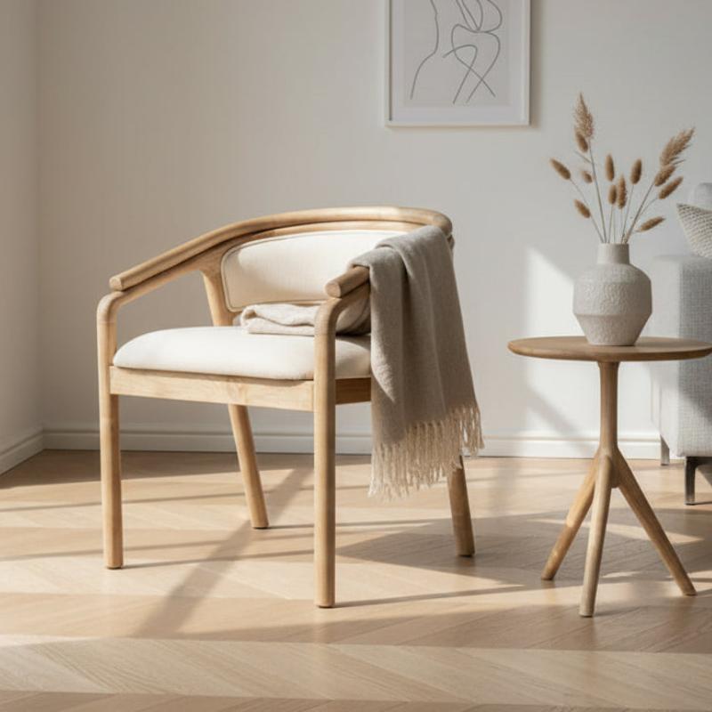 A light natural wood dining chair with cream upholstered seat and back, styled with a draped beige throw blanket, set in a bright minimalist room next to a small wooden side table.