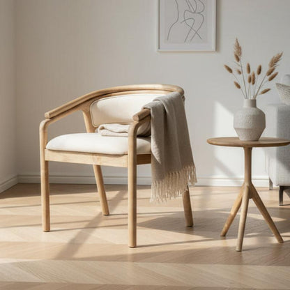 A light natural wood dining chair with cream upholstered seat and back, styled with a draped beige throw blanket, set in a bright minimalist room next to a small wooden side table.