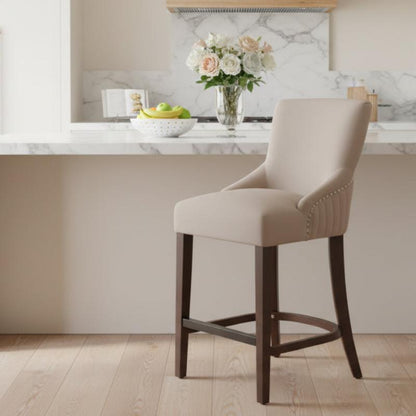A beige upholstered bar stool with dark wood legs and nailhead trim, positioned in front of a kitchen island with marble backsplash and countertop.