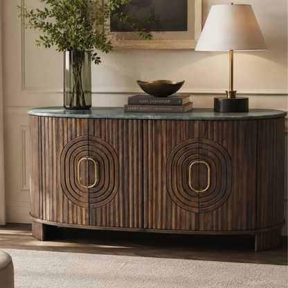 A curved wooden sideboard or credenza with vertical slats, two large oval recessed panels with gold-tone handles, and a green-blue stone top, styled in a living room with a lamp, books, a bowl, and a vase of greenery.