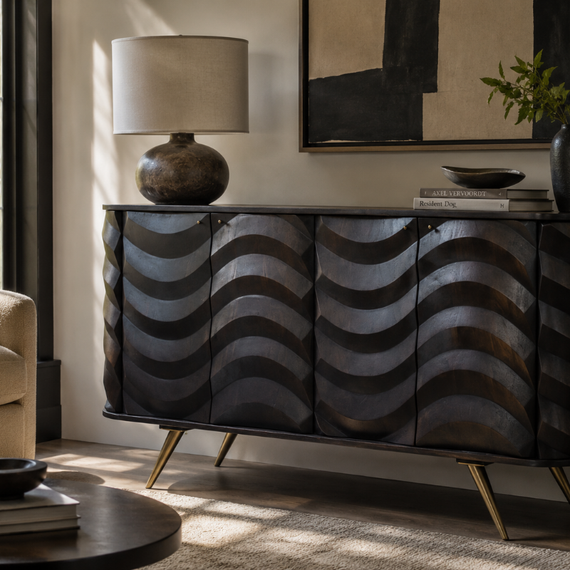 A modern sideboard or credenza with four front doors featuring a raised wavy pattern in dark metallic gray and black, supported by gold-toned angled legs. The cabinet is styled in a living room setting with a table lamp, books, and framed wall art.