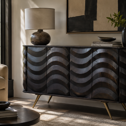 A modern sideboard or credenza with four front doors featuring a raised wavy pattern in dark metallic gray and black, supported by gold-toned angled legs. The cabinet is styled in a living room setting with a table lamp, books, and framed wall art.