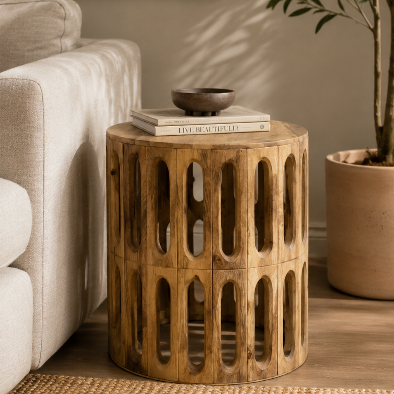 A round wooden end table with an open, lattice-like cylindrical design, styled in a warm living room next to a beige sofa and a potted plant.