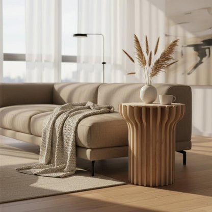 A modern beige fabric sofa in a bright, sunlit living room, styled with a textured cream throw blanket and a fluted wooden side table holding a white vase with dried grass and a small cup.
