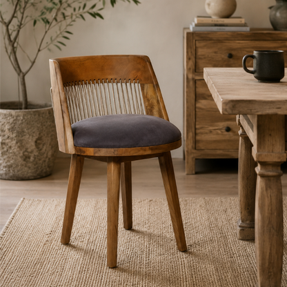 A wooden dining chair with a curved backrest, woven slat back, and a dark gray cushioned seat, shown in a rustic room next to a wooden table.
