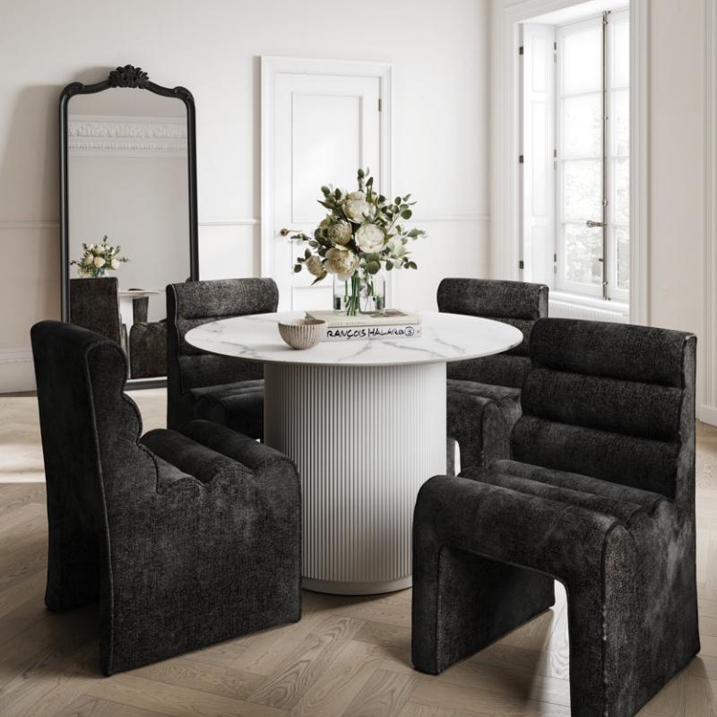 A dining set in a bright, classic room featuring a round white pedestal table and four dark gray upholstered baroque-style armchairs, with a large black-framed mirror on a light wood herringbone floor.