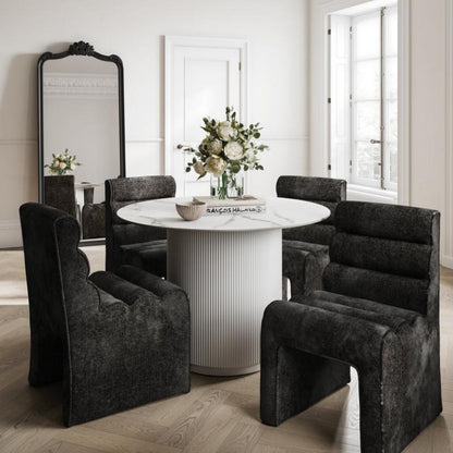 A dining set in a bright, classic room featuring a round white pedestal table and four dark gray upholstered baroque-style armchairs, with a large black-framed mirror on a light wood herringbone floor.