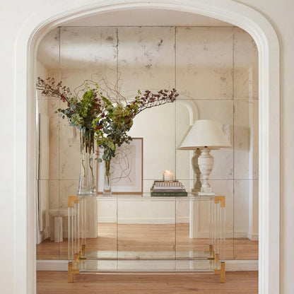 Acrylic console table with gold legs displaying a vase of flowers and a lamp in a room with wooden flooring.