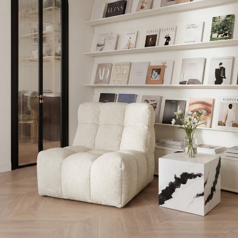 A cream-colored, tufted, low-profile lounge chair in a modern, minimalist room with light wood floors, white shelves, and a white marble-topped side table.