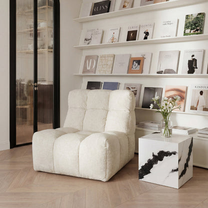 A cream-colored, tufted, low-profile lounge chair in a modern, minimalist room with light wood floors, white shelves, and a white marble-topped side table.