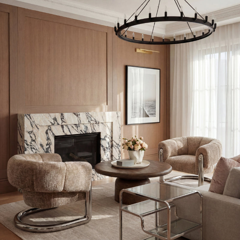 A luxury modern living room featuring two plush tufted boucle armchairs with chrome bases, a round wooden coffee table, a glass coffee cart, and a marble fireplace against a wood-paneled wall.