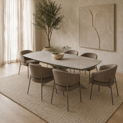 A modern dining set featuring a rectangular light gray stone-look table and six upholstered dining chairs in taupe, arranged on a large textured beige rug in a warm, minimalist dining room.