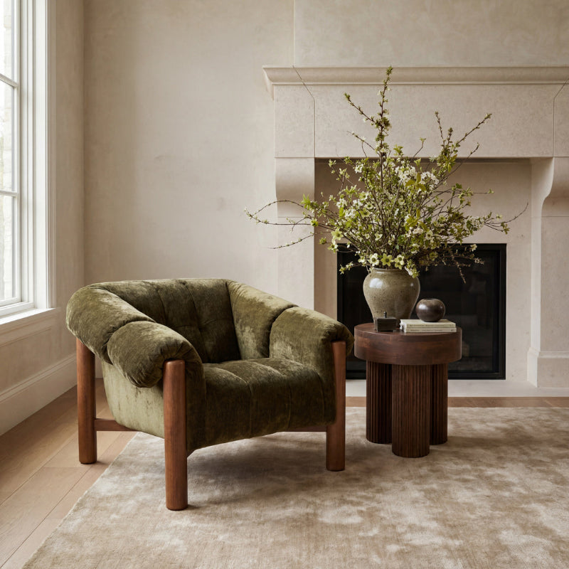 An olive green velvet armchair with wooden legs sits in a styled room next to a small round wooden side table holding a ceramic vase filled with branches.