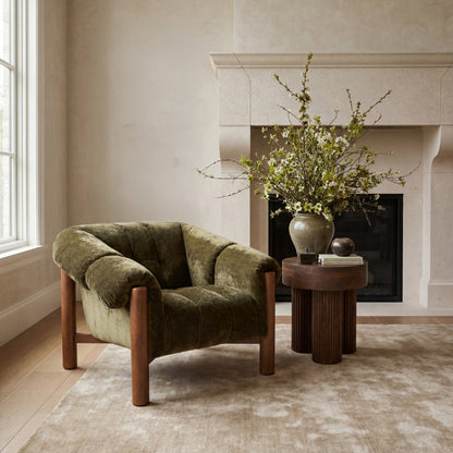 An olive green velvet armchair with wooden legs sits in a styled room next to a small round wooden side table holding a ceramic vase filled with branches.
