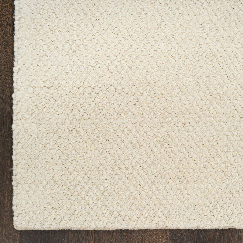 A close-up of a textured off-white or cream woven rug corner laid over a dark brown wooden floor.