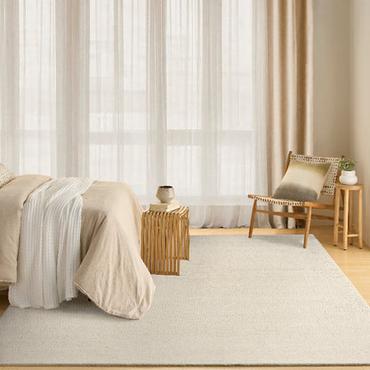 A large, light beige textured area rug in a bright, modern bedroom setting with a bed, wooden side table, and a chair by sheer white curtains.