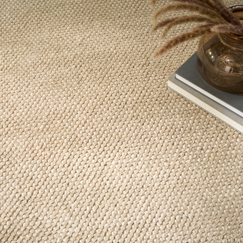 A close-up of a textured woven carpet or rug in a warm cream-beige tone, with a small stack of books and a brown glass vase with dried stems in the upper-right corner.