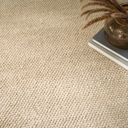 A close-up of a textured woven carpet or rug in a warm cream-beige tone, with a small stack of books and a brown glass vase with dried stems in the upper-right corner.