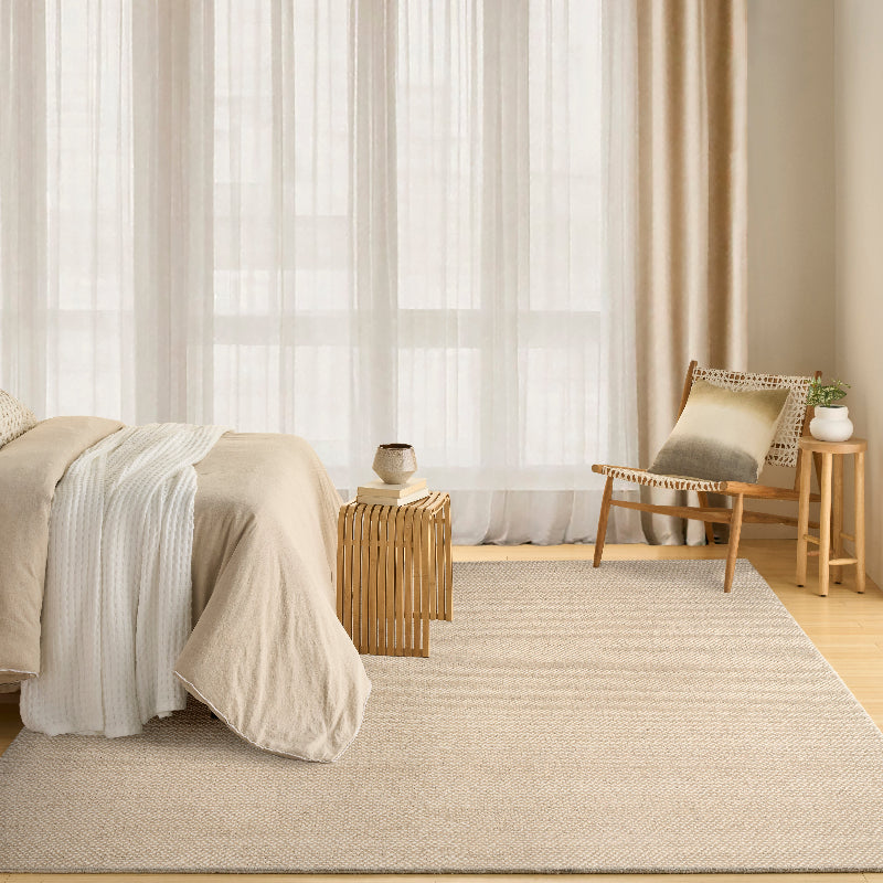 A large beige woven area rug in a bright, modern bedroom setting with a bed, wooden side table, and a chair with a cushion.