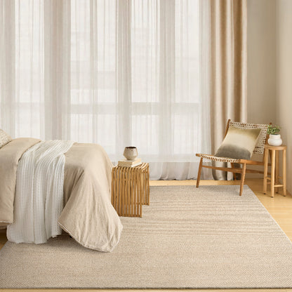 A large beige woven area rug in a bright, modern bedroom setting with a bed, wooden side table, and a chair with a cushion.