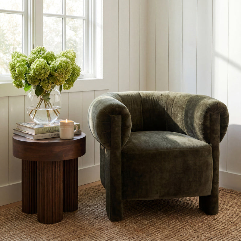 A dark olive green velvet armchair in a bright, sunlit room next to a round wooden side table with a vase of green hydrangeas, books, and a candle.