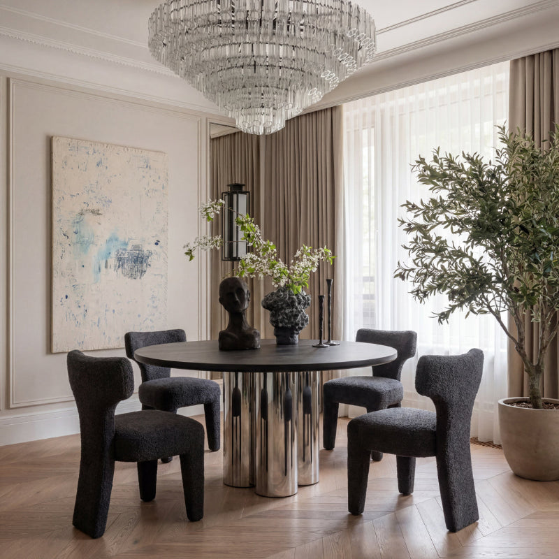 A luxury dining room featuring a round black table with a polished chrome pedestal base, surrounded by four dark gray upholstered sculptural chairs. The room is decorated with a large crystal chandelier, beige walls, a large abstract painting, and a potted olive tree.