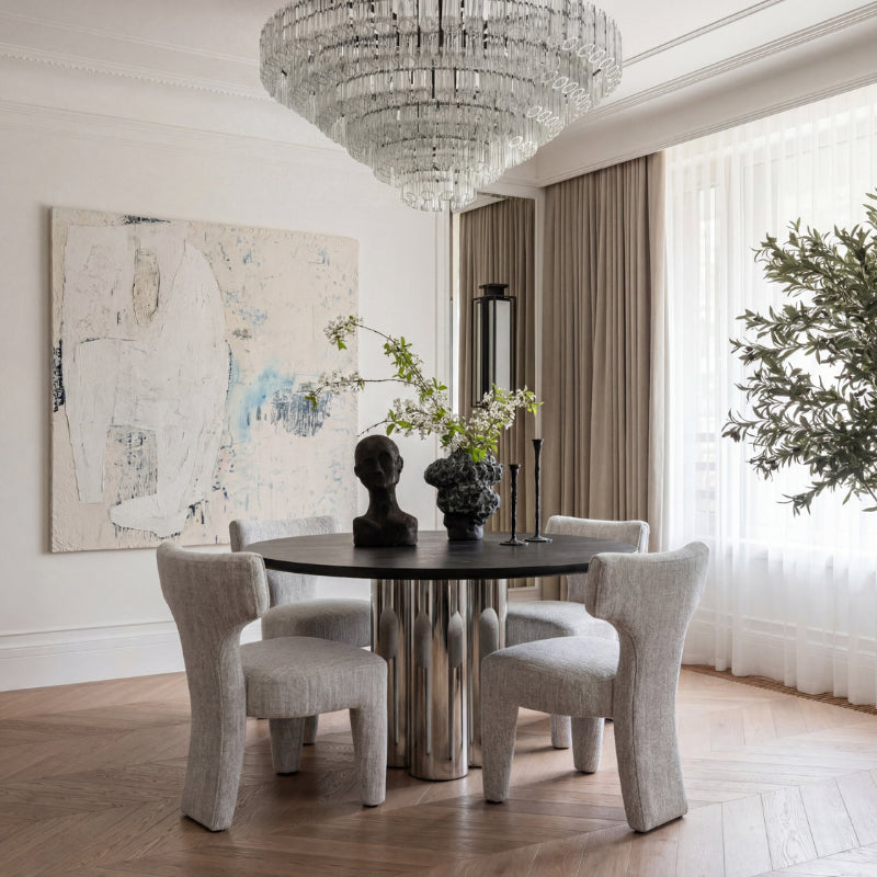 A modern dining room featuring a round black-top table with four light beige upholstered chairs, a large crystal chandelier, beige curtains, and a large abstract painting on the wall.