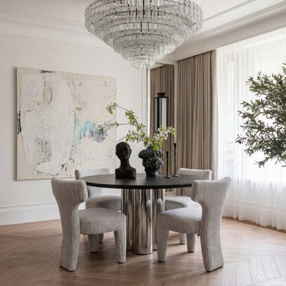 A modern dining room featuring a round black-top table with four light beige upholstered chairs, a large crystal chandelier, beige curtains, and a large abstract painting on the wall.