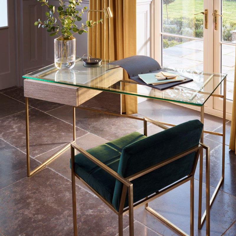 Modern glass desk with gold frame and green chair in a room with large windows.