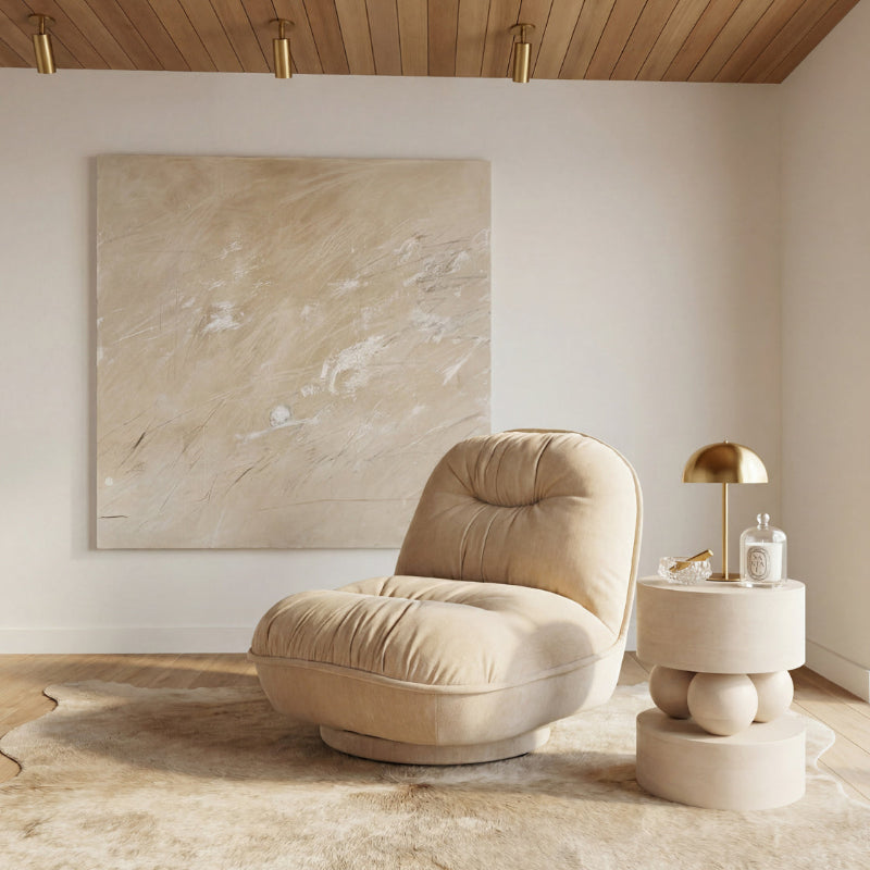 A plush tufted beige lounge chair sits in a styled interior room with a large abstract canvas on the wall, a wood ceiling, and a small round side table with a gold lamp and glass bottle.