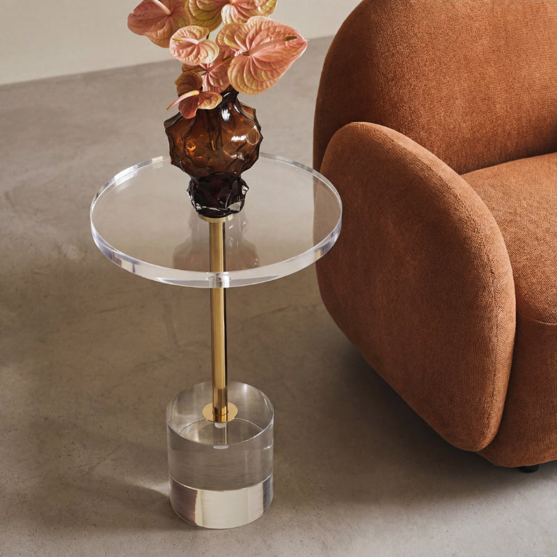 Clear glass side table with gold accent next to a brown armchair on a neutral background