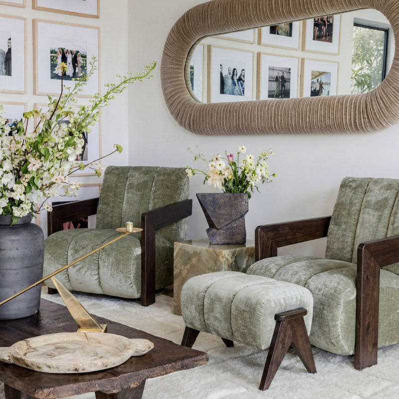 A styled living room featuring two sage green velvet armchairs with dark wood frames and a matching ottoman, set on a light rug beneath a large textured oval mirror.