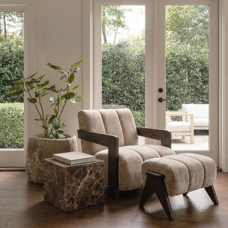 A modern upholstered lounge chair with a matching foot stool in a warm beige fabric and dark wood, styled in a bright living room with a stone planter, marble side table, and glass doors.